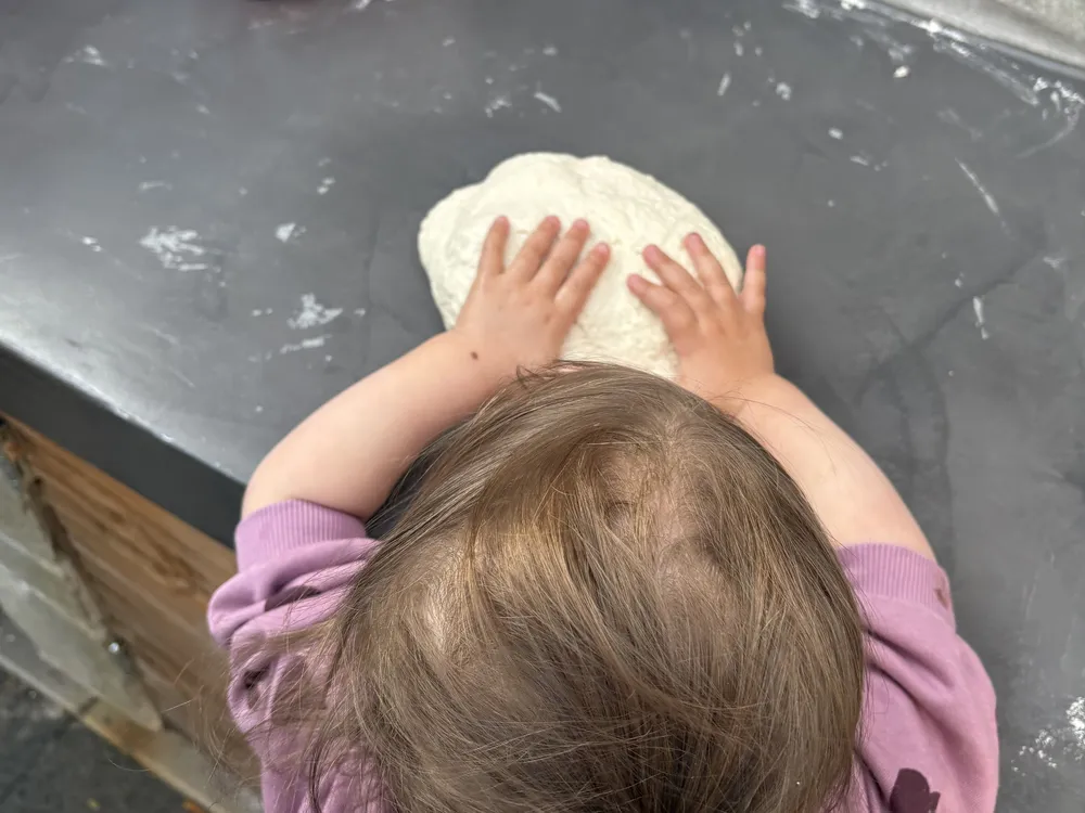 Een kind drukt met beide handen een bol pizzadeeg plat op een grijs werkblad, met een pak bloem op de achtergrond