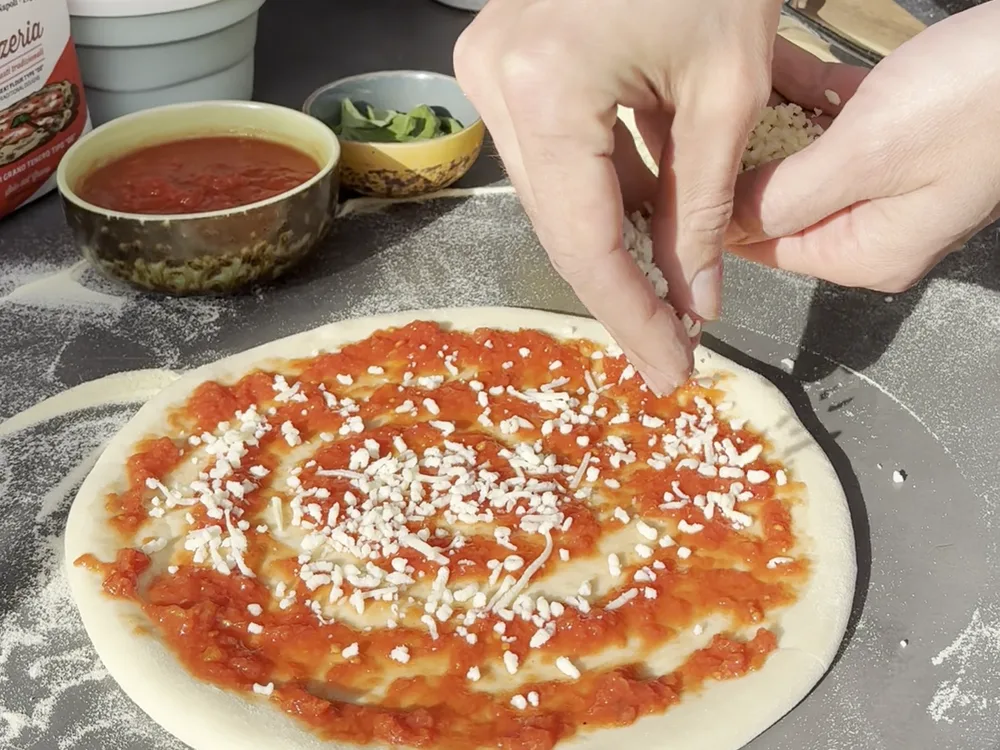 Handen die mozzarella over een pizzabodem met tomatensaus strooien op een buitenwerkblad, met een zak bloem en bakjes op de achtergrond