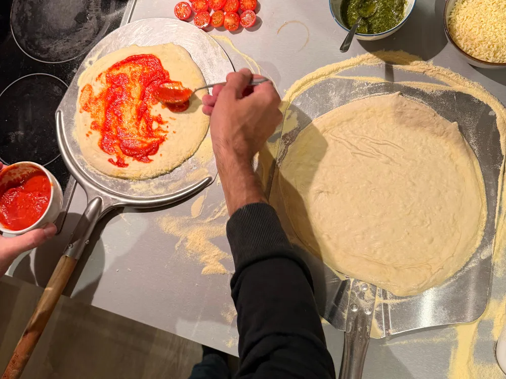 Iemand smeert tomatensaus op een pizzabodem aan het aanrecht, met bakjes cherrytomaten, pesto en geraspte kaas ernaast
