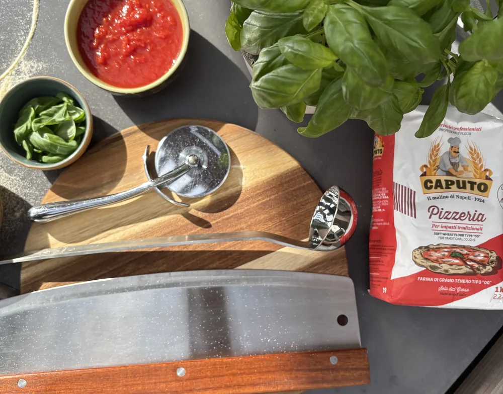 Pizzabereiding van bovenaf op een donker werkblad: een ronde houten plank met pizzawiel, lepel en wiegmess, naast een kom tomatensaus, vers basilicum en een zak bloem