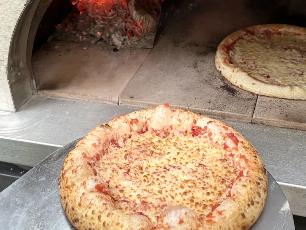 Gebakken margherita op een pizzaschep voor de houtoven, met een tweede pizza en gloeiende kolen in de oven