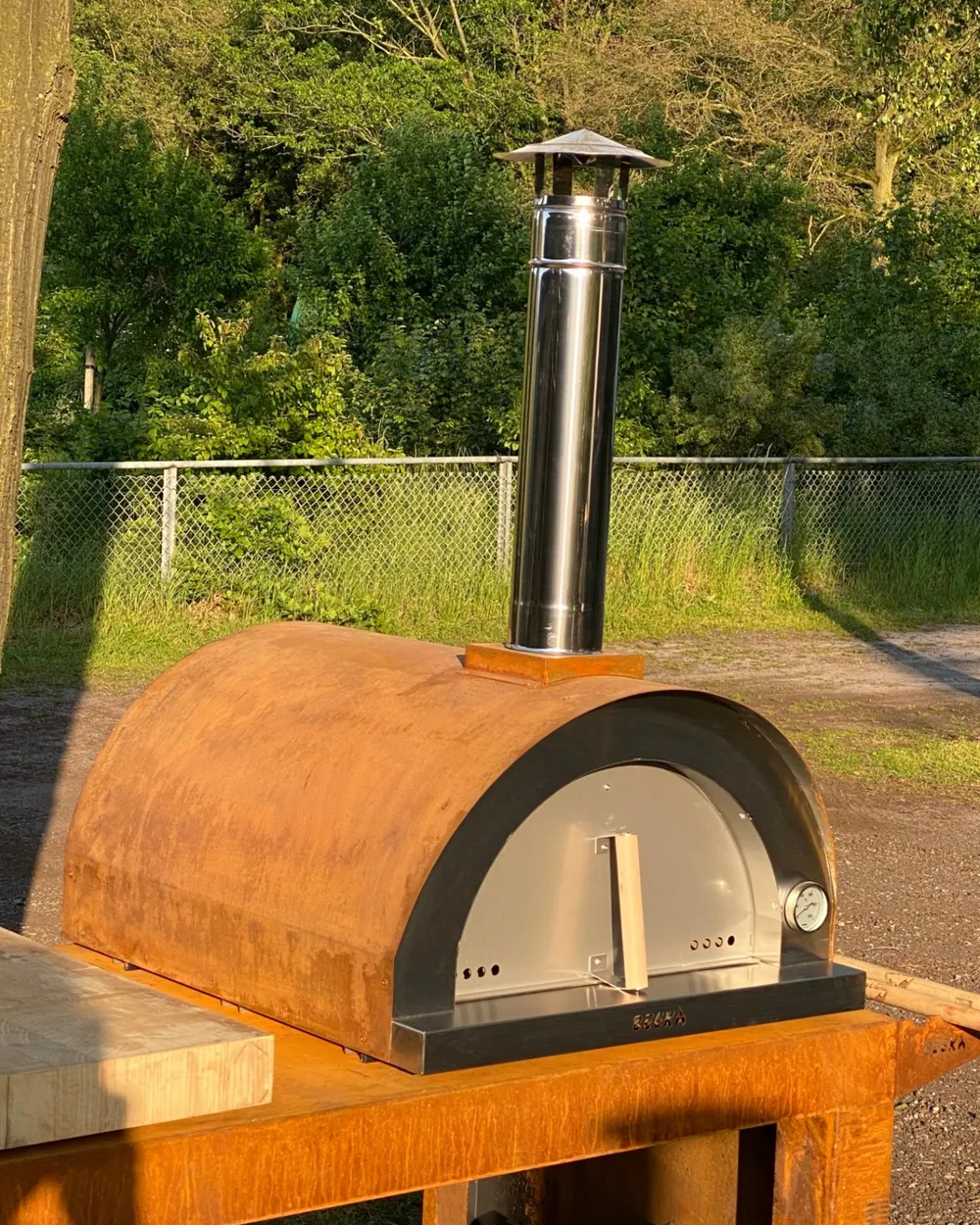 Houtgestookte pizzaoven met roestbruine koepel en schoorsteen buiten op een onderstel, met groen op de achtergrond