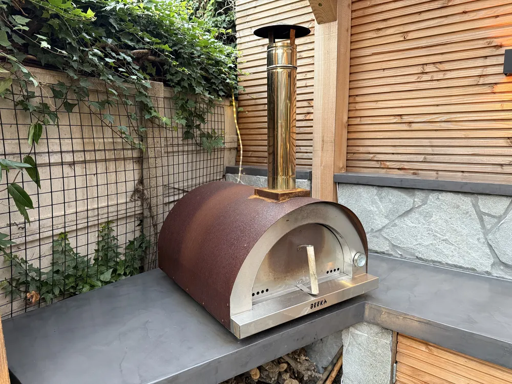 Pizzaoven met koperkleurige koepel op een betonnen werkblad in de tuin, tegen een houten schutting met klimop