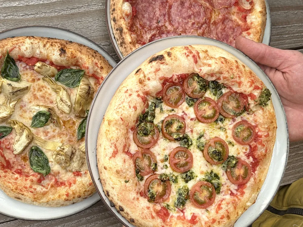 Drie vers gebakken pizza's op borden op een houten tuintafel, een ervan wordt vastgehouden