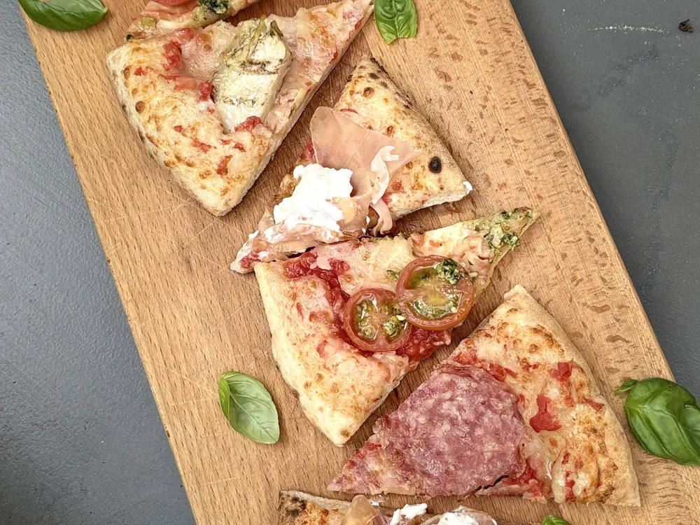 Pizzapunten met artisjok, parmaham en salami op een lange houten plank, met basilie eromheen